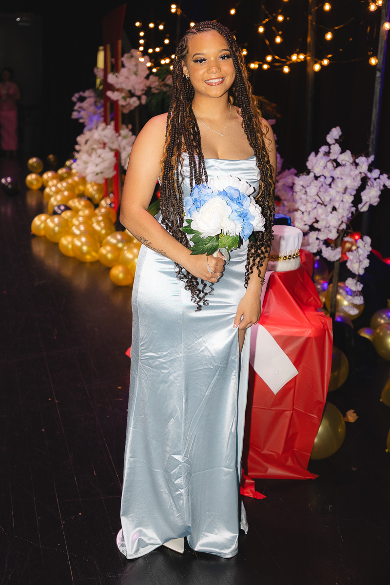 Young woman in a silver strapless gown holding a blue-and-white bouquet at a festive indoor event with warm lights and balloons in the background.
