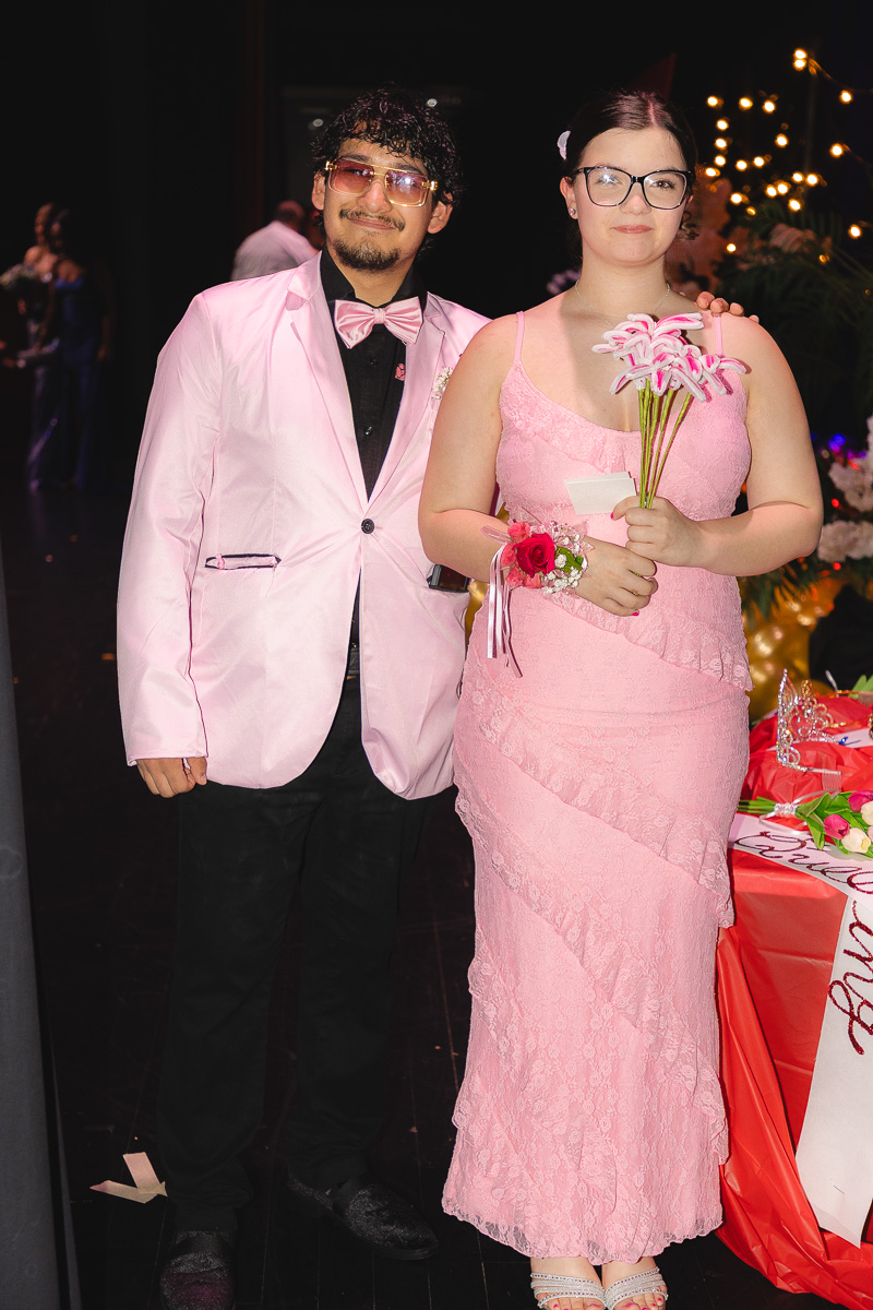 A couple posing at a formal event: man in a pink blazer and bow tie beside a woman in a pink lace dress holding pink lilies and a corsage.