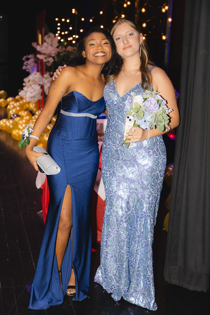 Two young women in elegant blue gowns pose together on a decorated event stage, one holding a clutch and corsage.