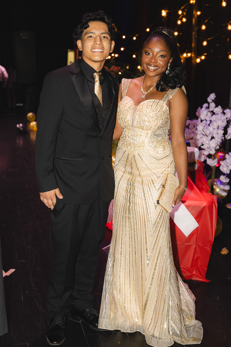 Couple posing at a formal event: man in a black suit with gold tie and woman in a gold beaded gown holding a gold clutch, standing under string lights.