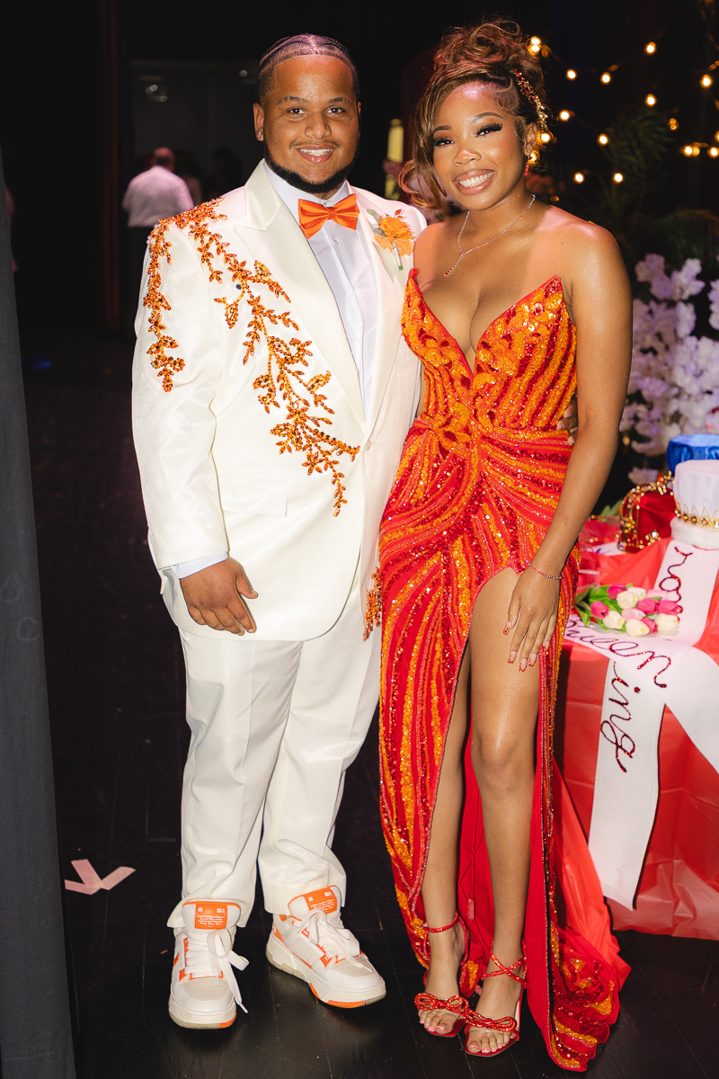 Couple posing at a formal event: man in a white suit with orange embroidery and bow tie, woman in a red embellished gown with a high slit.