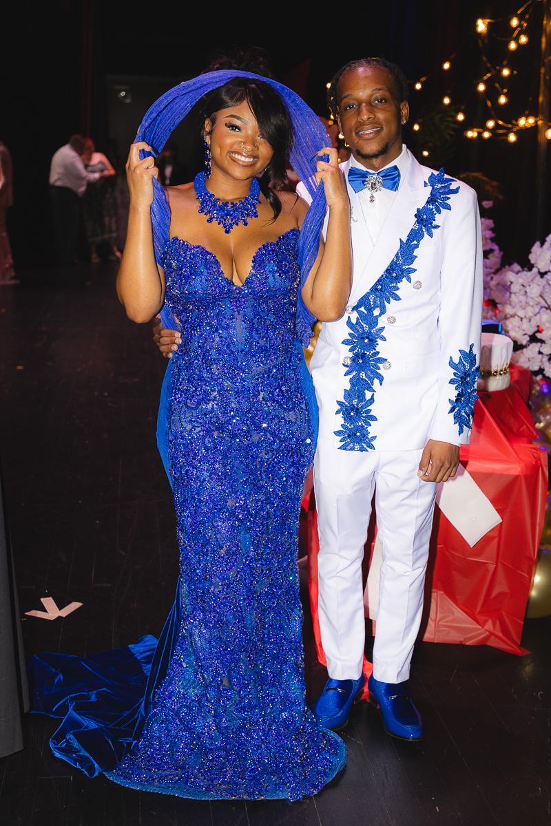 Couple posing at a formal event: woman in a sparkling royal blue gown with matching head wrap and necklace, man in a white suit with blue embroidery, both smiling under string lights