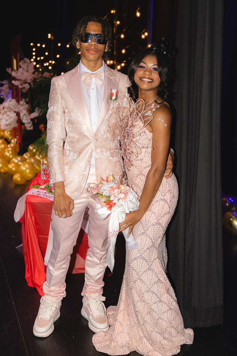 Couple dressed in coordinated pink formalwear posing together at a decorated event, the man in a pink suit and bow tie, the woman in a bead‑embellished pink gown holding a bouquet.