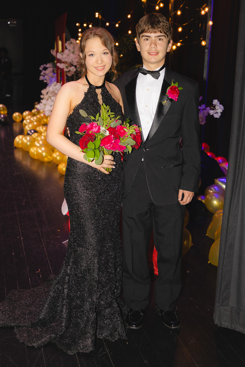 Young couple in formal wear posing on a decorated stage; woman holds a bouquet of red roses, man wears a black tuxedo with a red boutonniere.