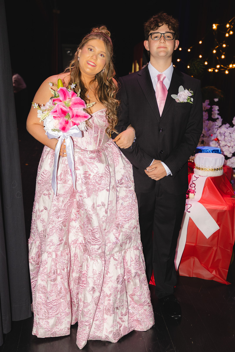 Prom couple posing together: girl in a pink lace ball gown holding a bouquet, boy in a black suit with a pink tie.