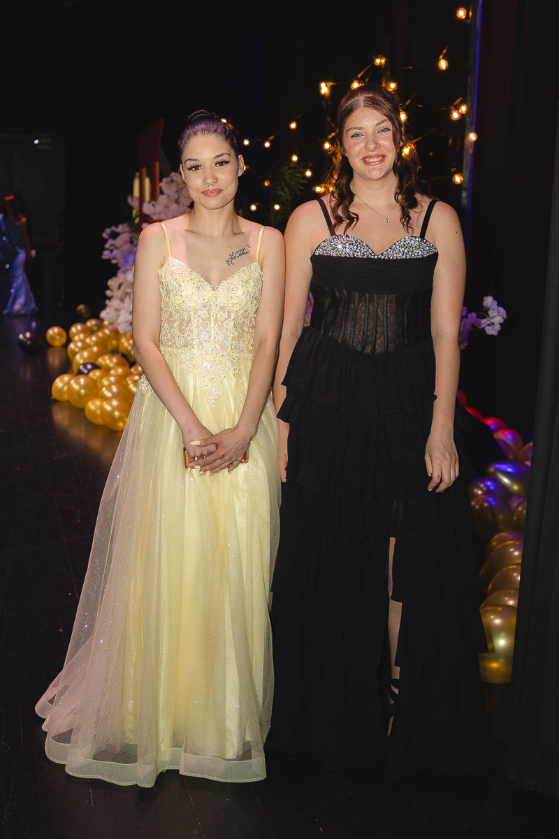 Two young women in formal gowns posing together at an event, with string lights and balloons in the background.