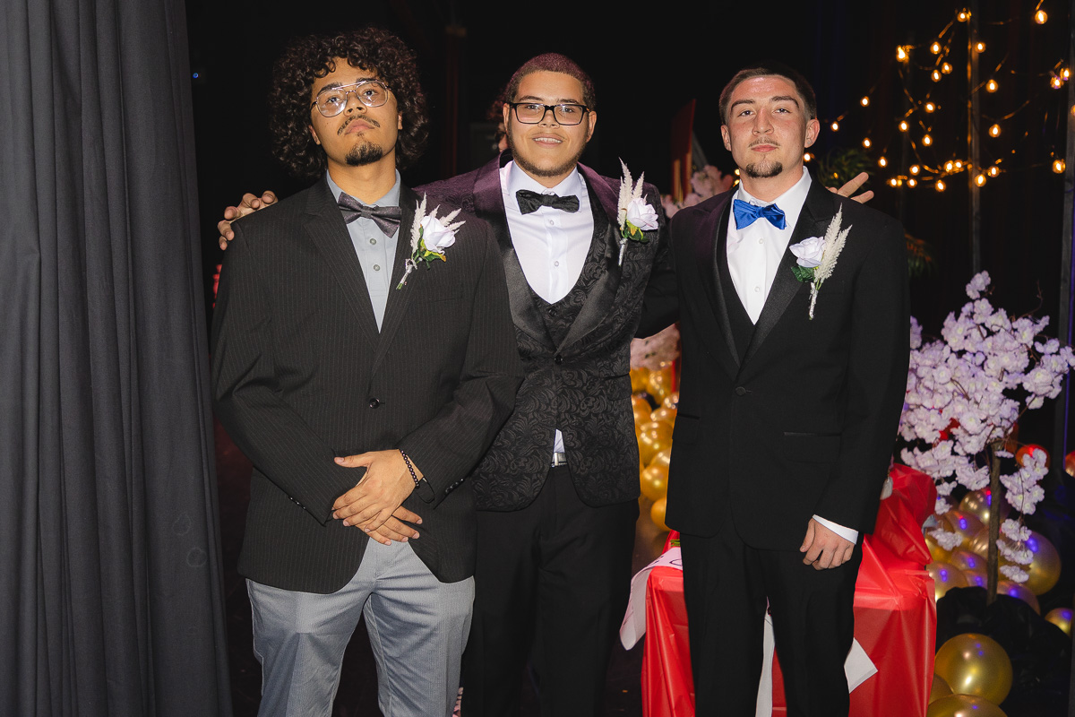 Three young men in tuxedos pose together at a decorated formal event, each with a boutonniere on the lapel, smiling for the camera.