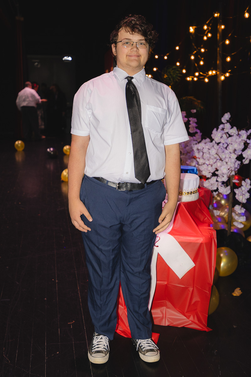 Young man in a white shirt and black tie, navy pants, and sneakers stands at a party table with gift bags and string lights in the background.