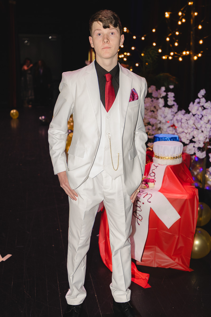 Young man in a white suit with a red tie posing at a festive event with string lights in the background.