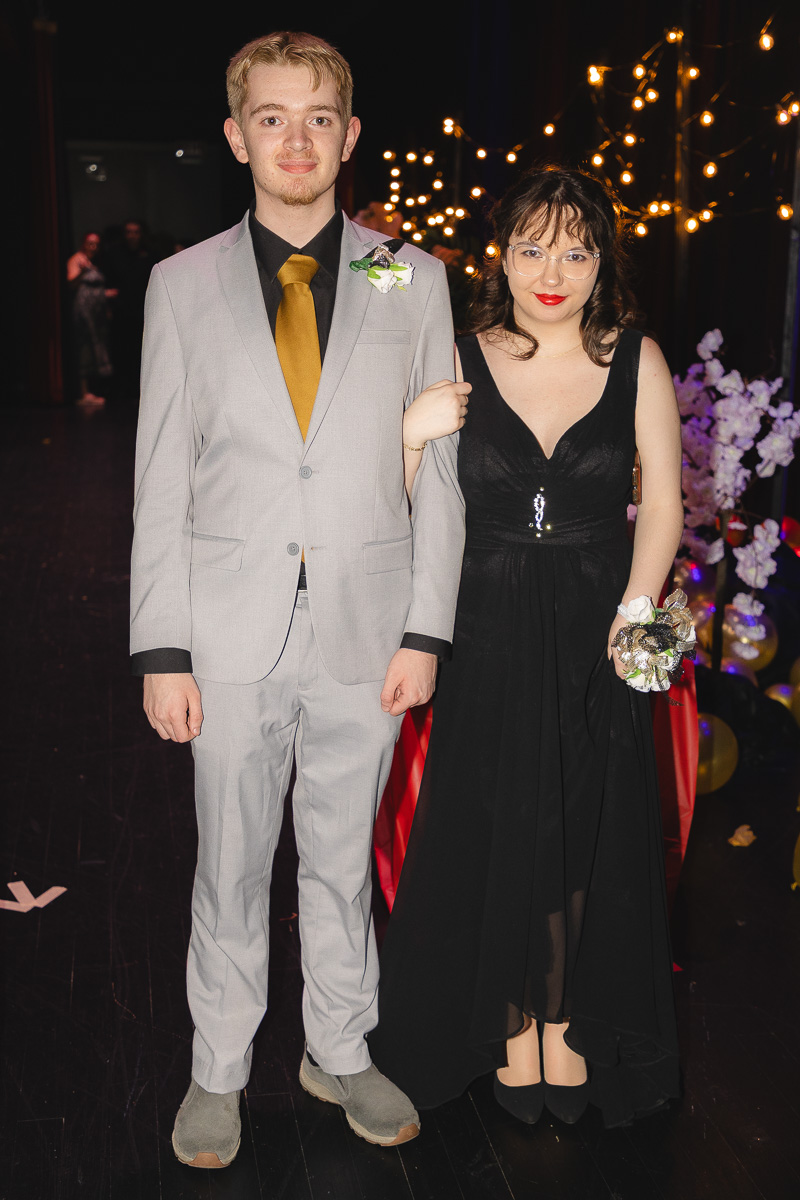 Couple in formal wear posing at a gala under string lights; man in gray suit with mustard tie and boutonniere, woman in black evening gown with corsage.