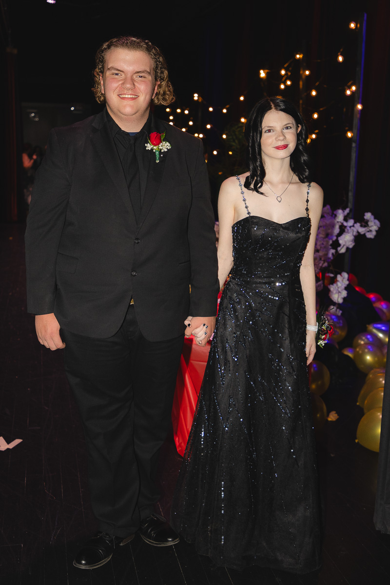 Couple in formal attire posing and holding hands at a gala with warm string lights in the background.