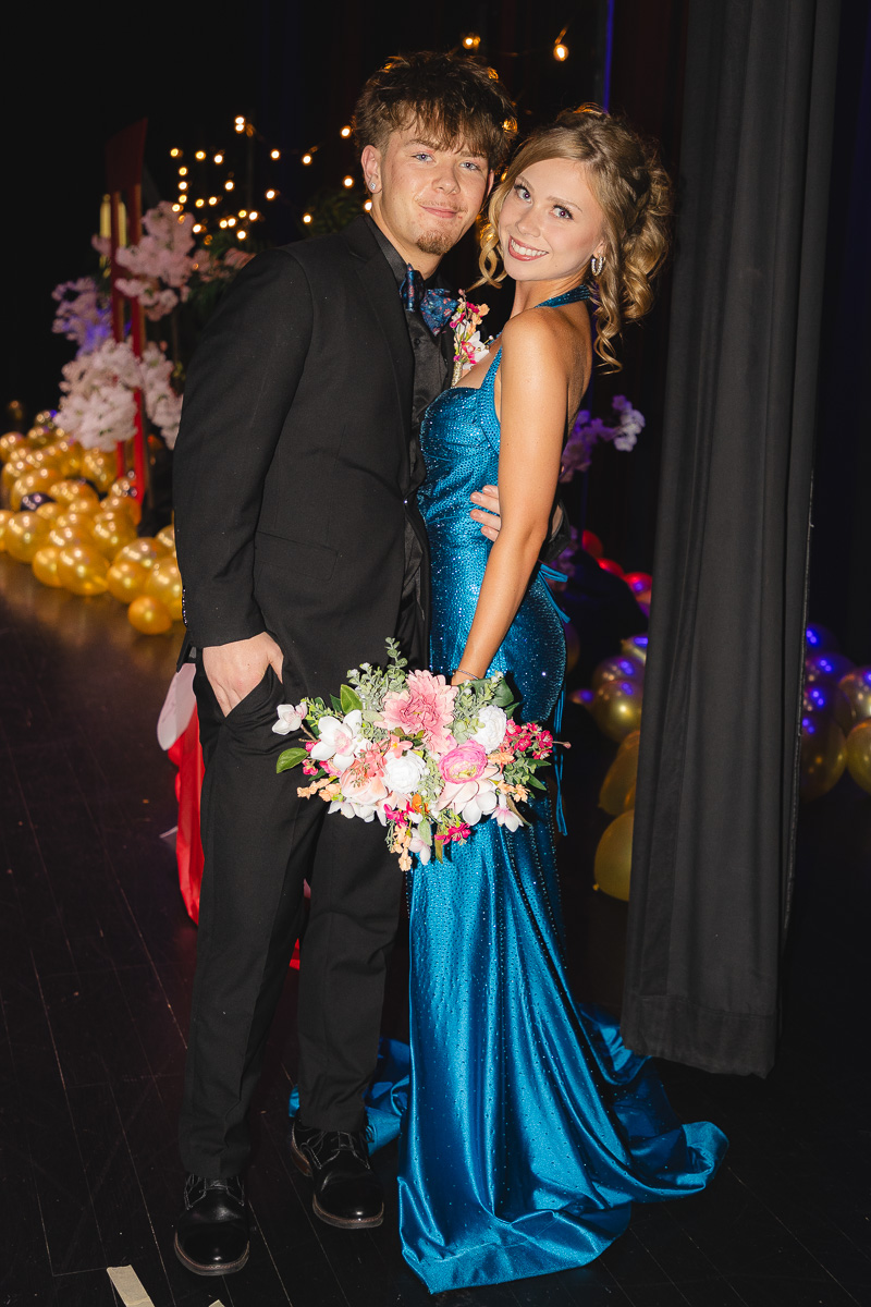 Smiling couple poses backstage at a formal event: man in a black suit, woman in a sparkly blue gown holding a pink‑and‑white bouquet.