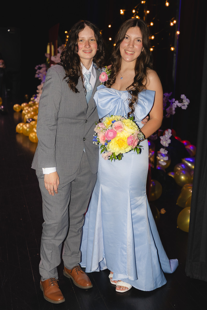 Two young people in formal attire posing on a decorated stage; woman in light blue gown holds bouquet.