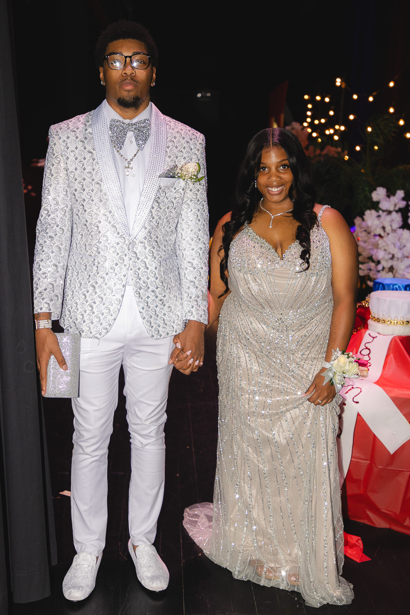 A couple in formal attire posing hand-in-hand at a party; man in a silver patterned suit and bow tie, woman in a sparkling silver gown.