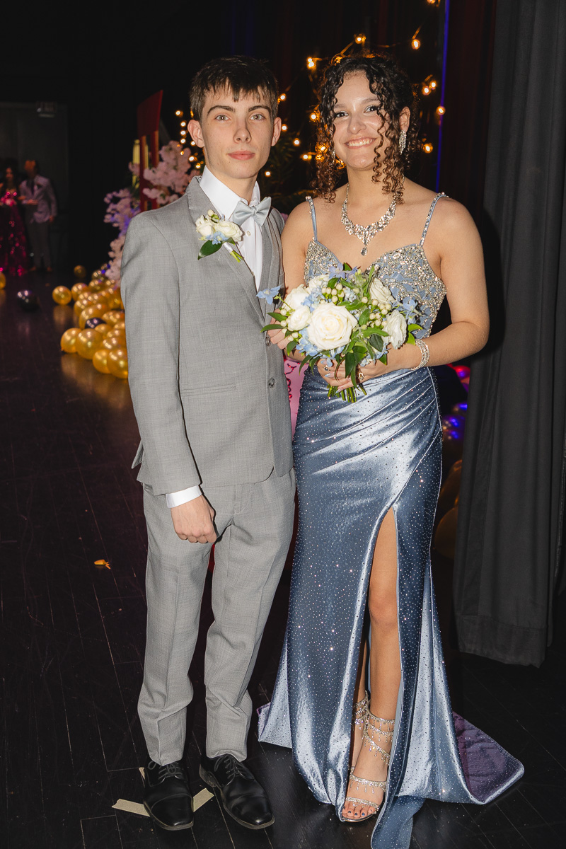 Young man in a gray suit with a white bow tie stands beside a woman in a glittery blue gown, both holding flowers at a formal event.
