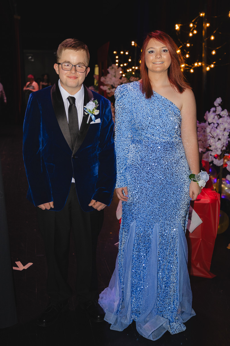 Young man in a blue velvet suit with boutonniere and tie, and a young woman in a one-shoulder blue sequin gown, posing at a formal event with warm string lights in the background.