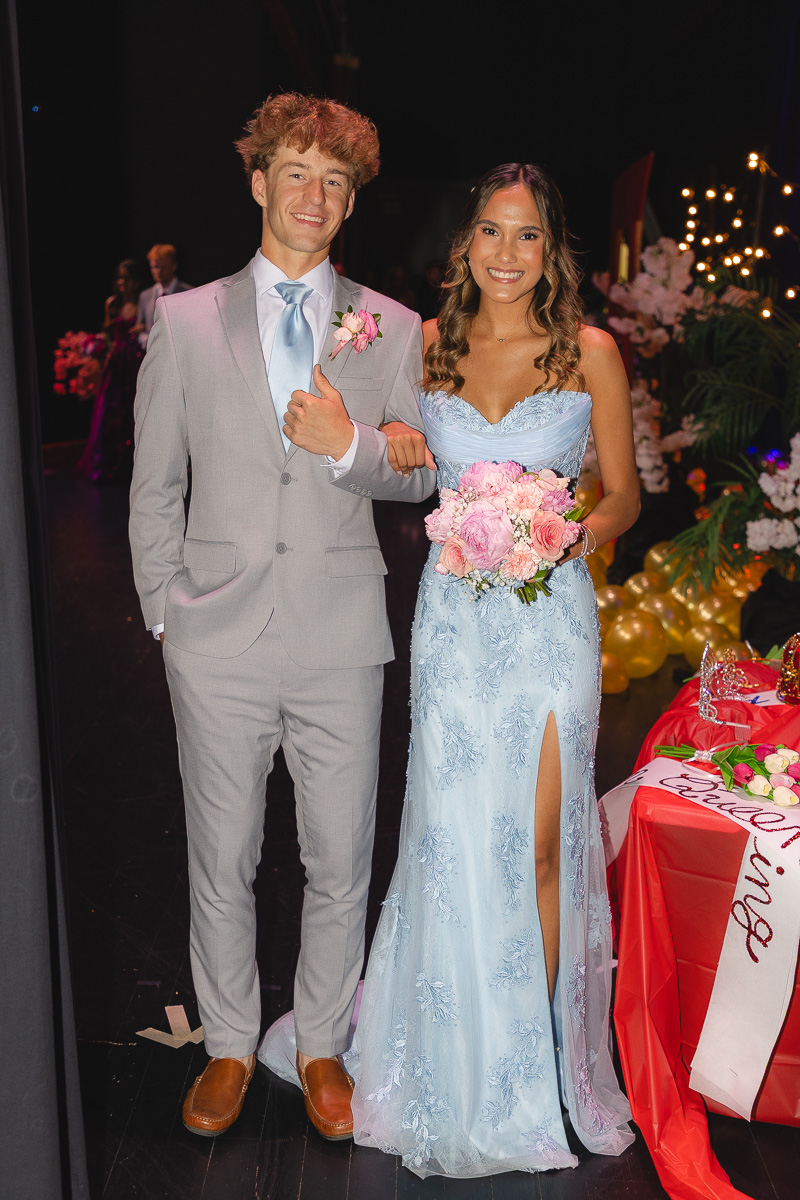 Couple in formal wear posing together at a ballroom event: man in light gray suit with a blue tie and pink boutonnière, woman in a light blue gown holding a pink bouquet.
