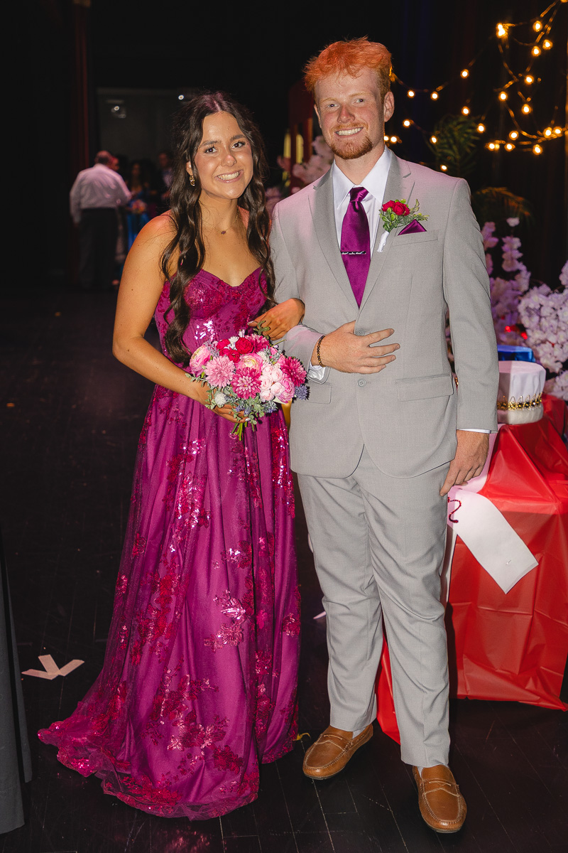 Couple posing at a formal event; woman in a magenta sequined gown holding a pink bouquet, man in a gray suit with a purple tie.