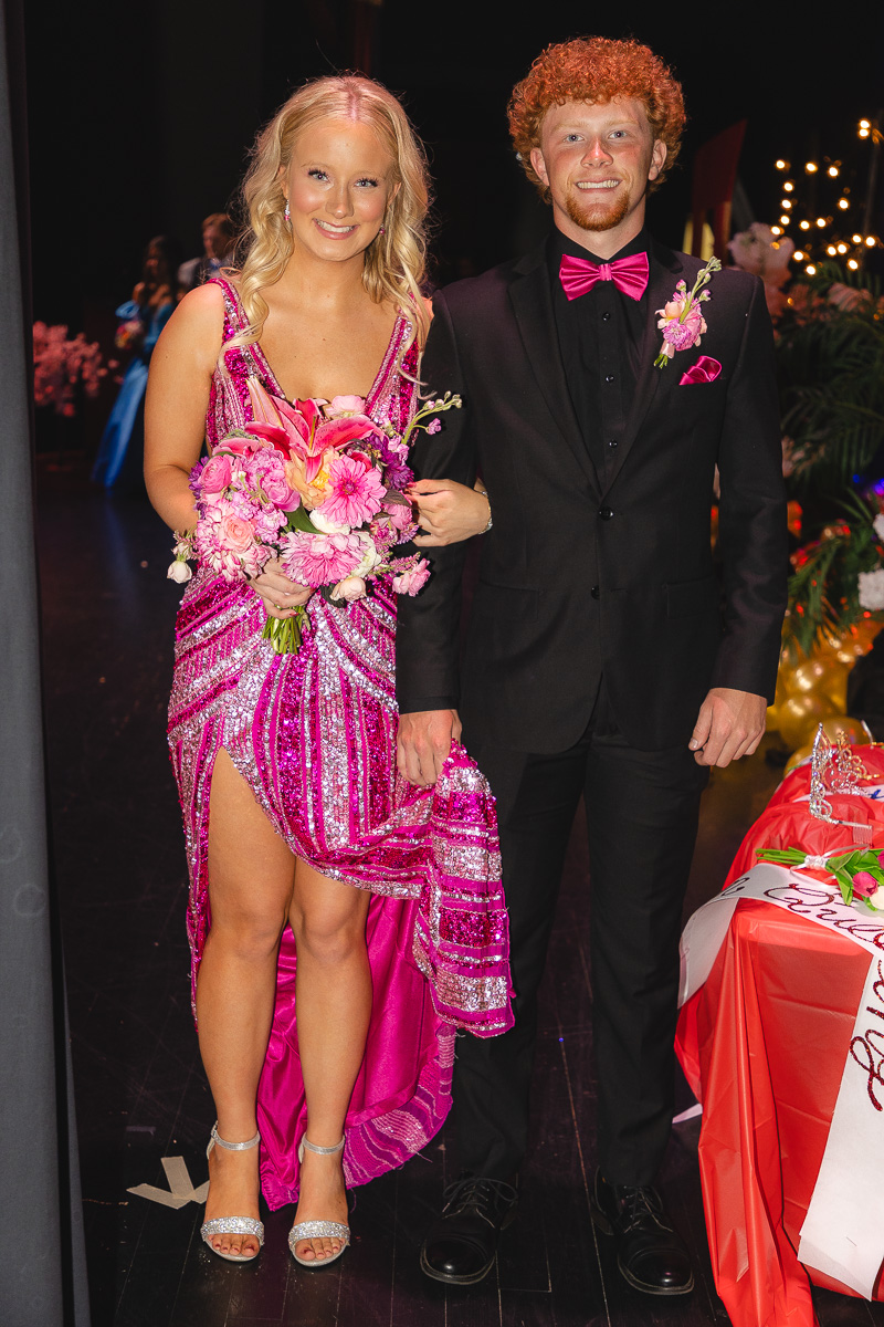 Couple walking backstage at a formal event: woman in a pink sequined gown holding a bouquet, man in a black suit with pink bow tie and boutonniere.