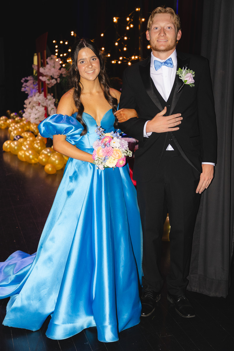 Woman in a bright blue ball gown holding a bouquet, standing beside a man in a black tuxedo with a light blue bow tie.