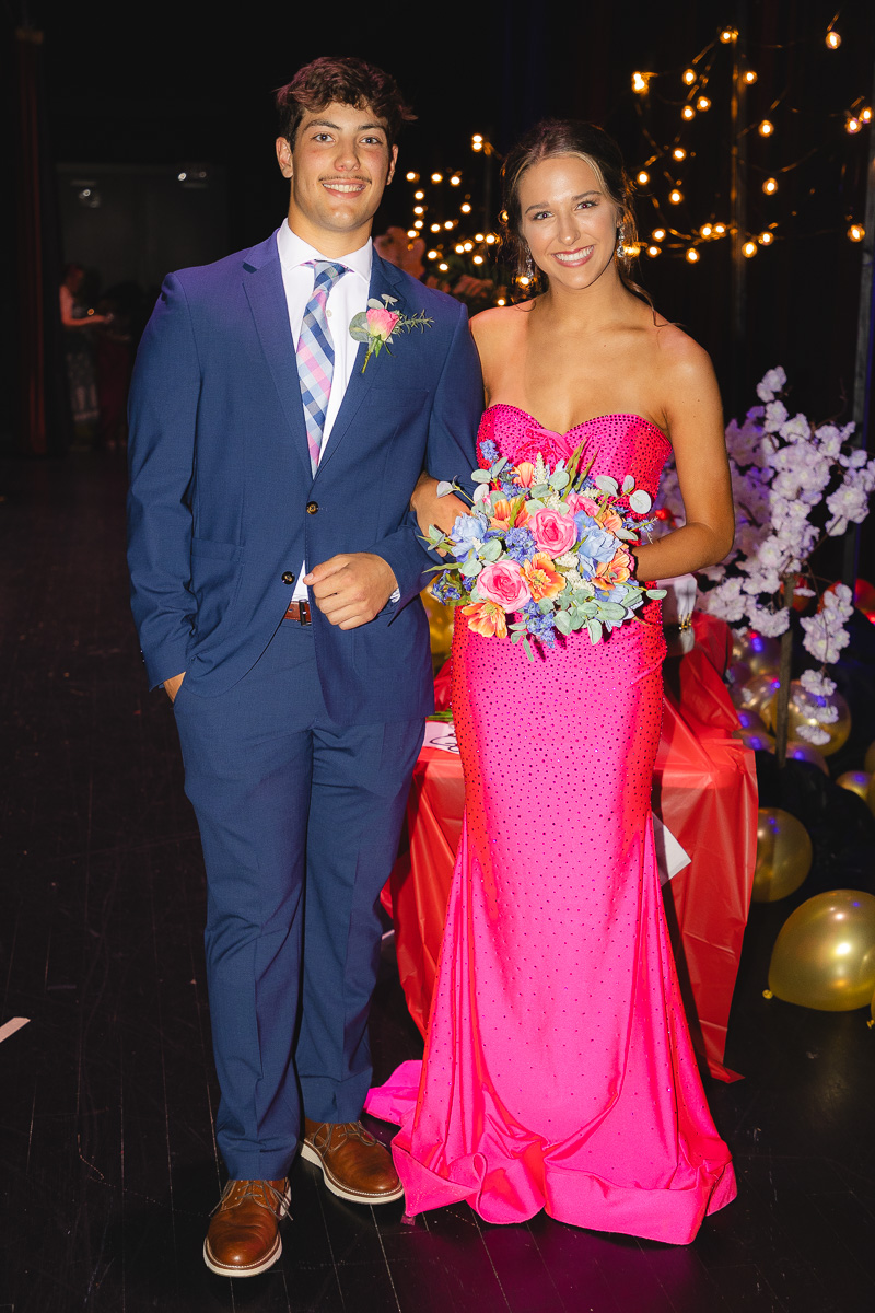 Couple posing at a formal event: man in a blue suit with boutonniere and a pink plaid tie, woman in a bright pink strapless gown holding a colorful bouquet, string lights behind them.