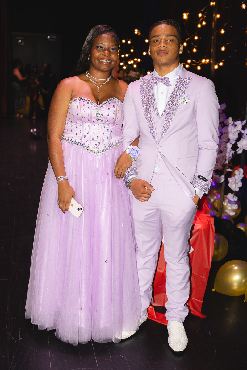 Young woman in a lavender ball gown and a young man in a lavender suit pose arm-in-arm at a festively lit formal event, smiling.