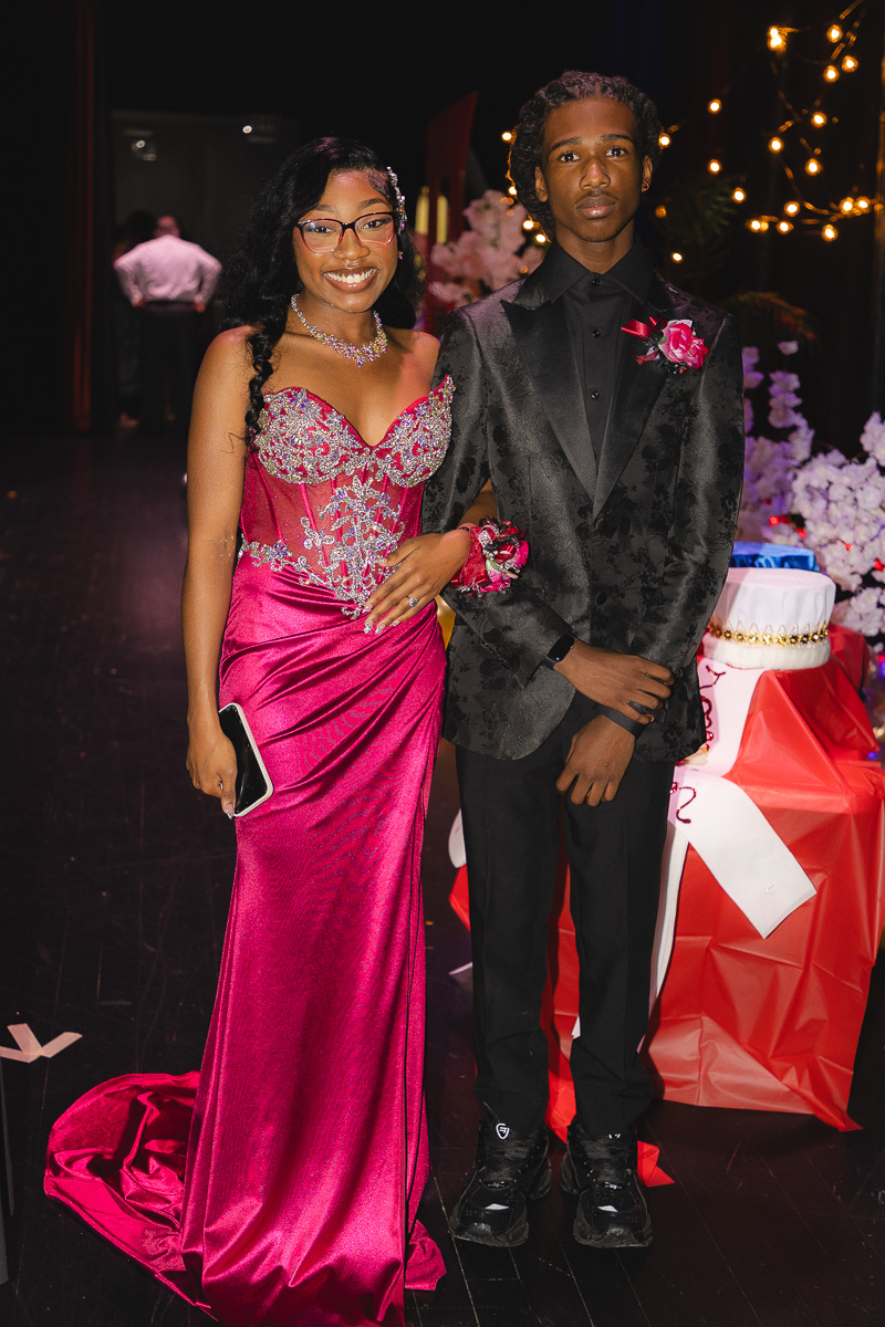 Young couple in formal attire posing for a photo at a decorated event; the girl wears a magenta bejeweled gown and necklace, holding a small clutch, while the boy wears a black suit with a pink boutonniere and corsage.