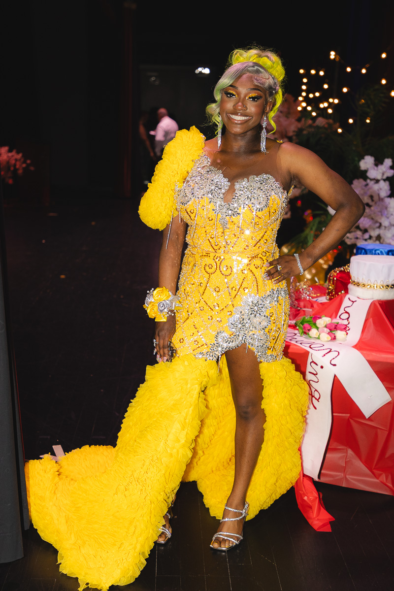 Woman in a bright yellow embellished gown with a dramatic feathered train posing at a gala, smiling confidently.