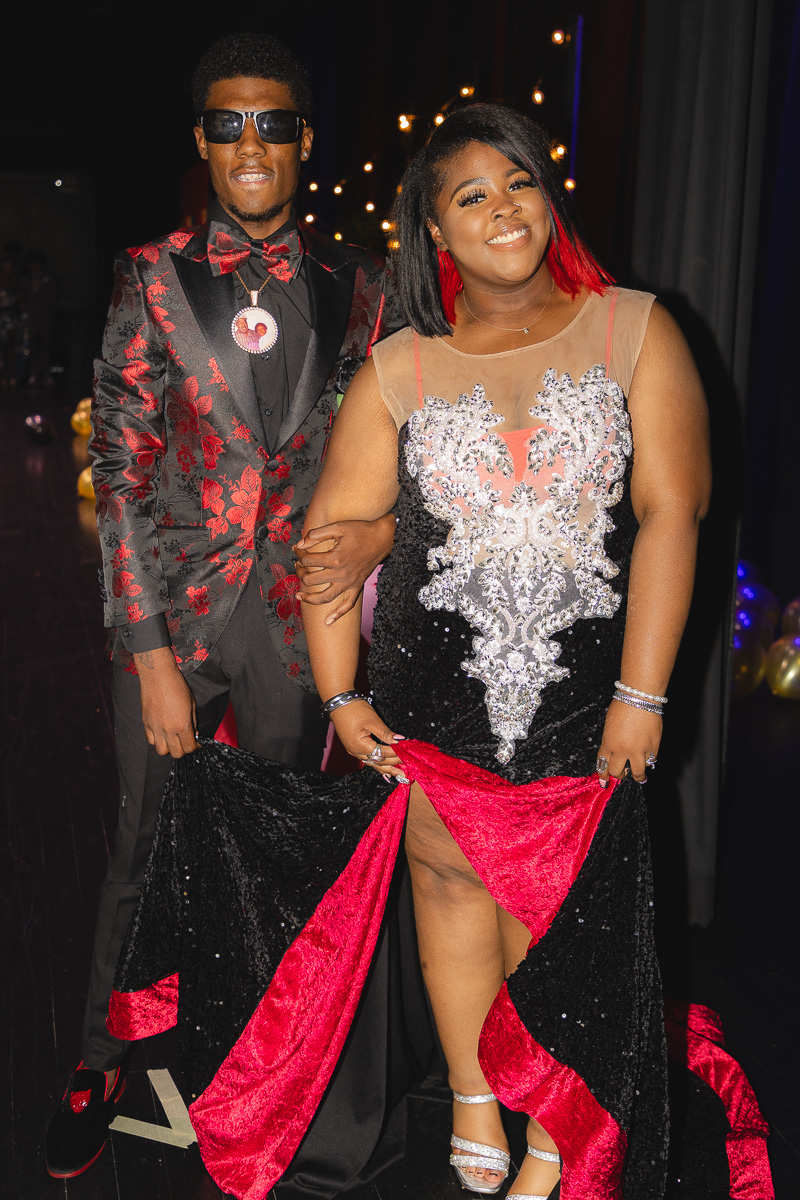 Man in a black suit with red floral pattern and a bow tie stands beside a smiling woman in a jeweled black-and-red gown at a formal event, with lights in the background.
