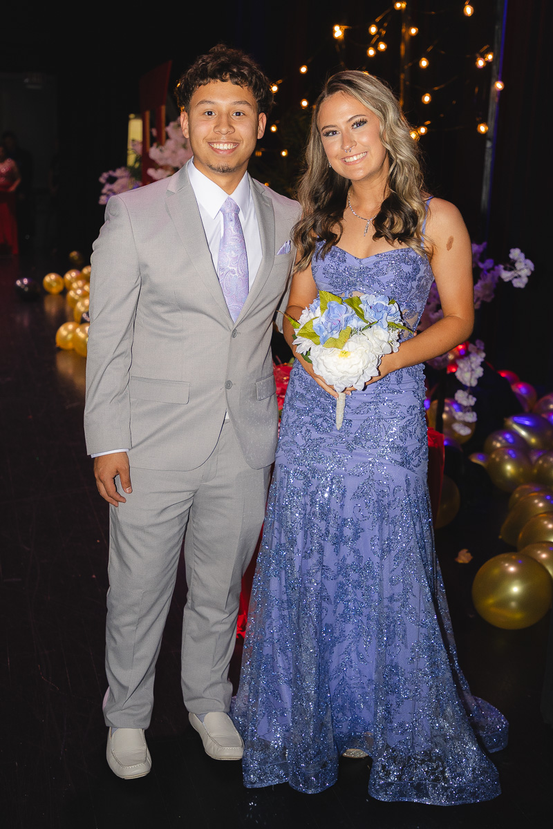 Young couple posing at a formal event: man in a light gray suit, woman in a blue sequined gown holding a bouquet, smiling under string lights and balloons.