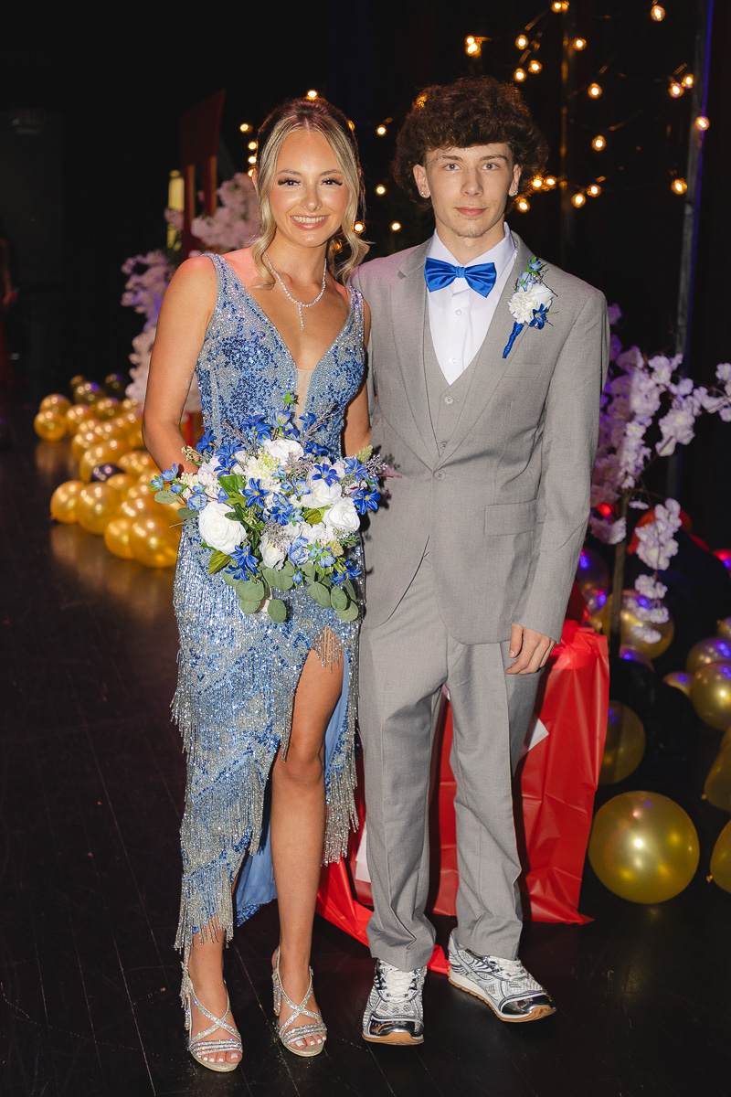 Couple posing at a formal event: woman in a blue sequined gown holding a white-blue bouquet, man in a light gray suit with a blue bow tie, background of string lights and balloons.