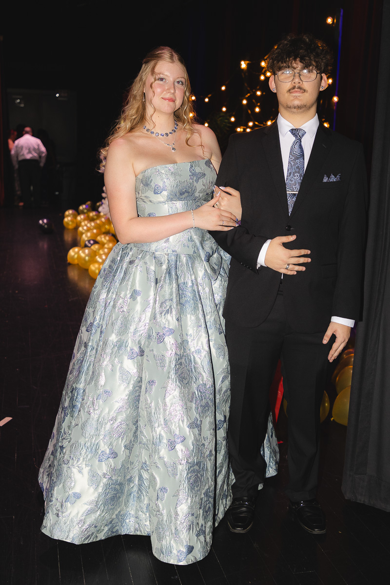 Couple posing at a formal event: woman in a silver-blue floral strapless gown and a man in a black suit with a patterned tie, arm in arm against a backdrop of string lights and balloons.