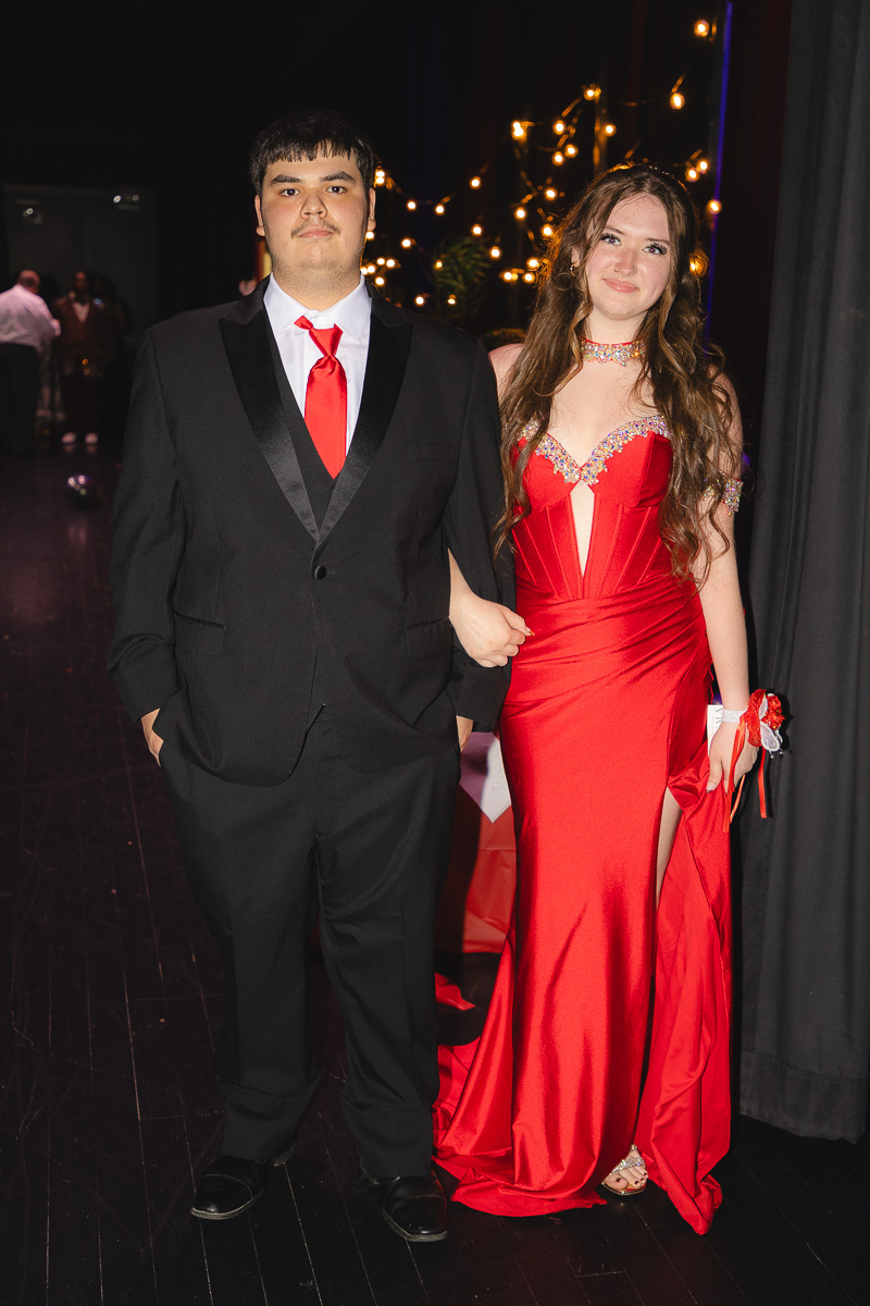 Young man in a black suit with a red tie and white shirt stands beside a young woman in a red, beaded evening gown at a formal event; string lights glow in the background as they walk down a stage or hallway, arms linked.