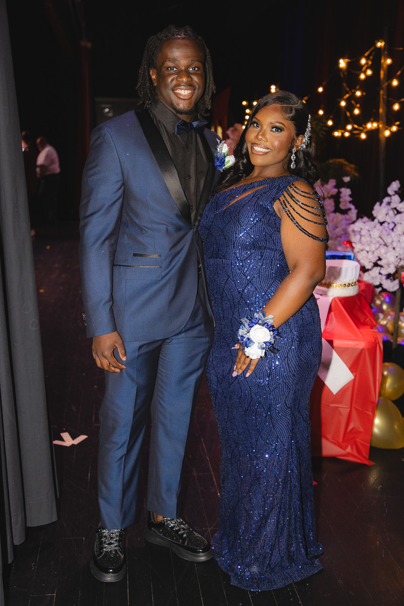 Smiling couple in formal blue attire posing at a decorated party with string lights in the background.