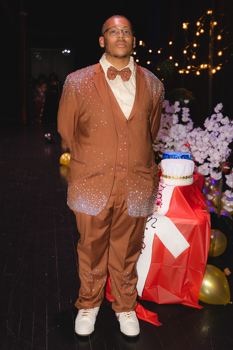 Man in a brown sequined suit and bow tie posing at a festive party with string lights and wrapped gifts nearby.