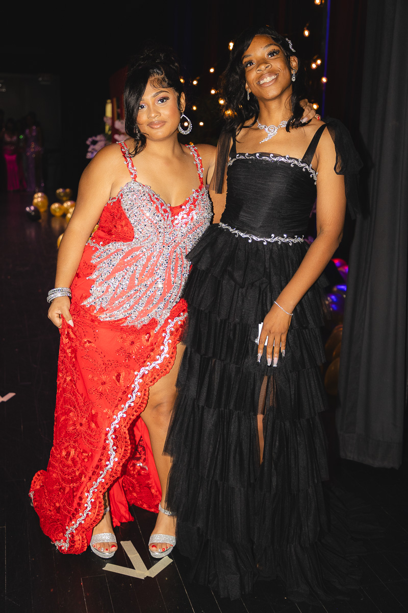 Two young women in formal gowns posing together at a nighttime event with string lights behind them.