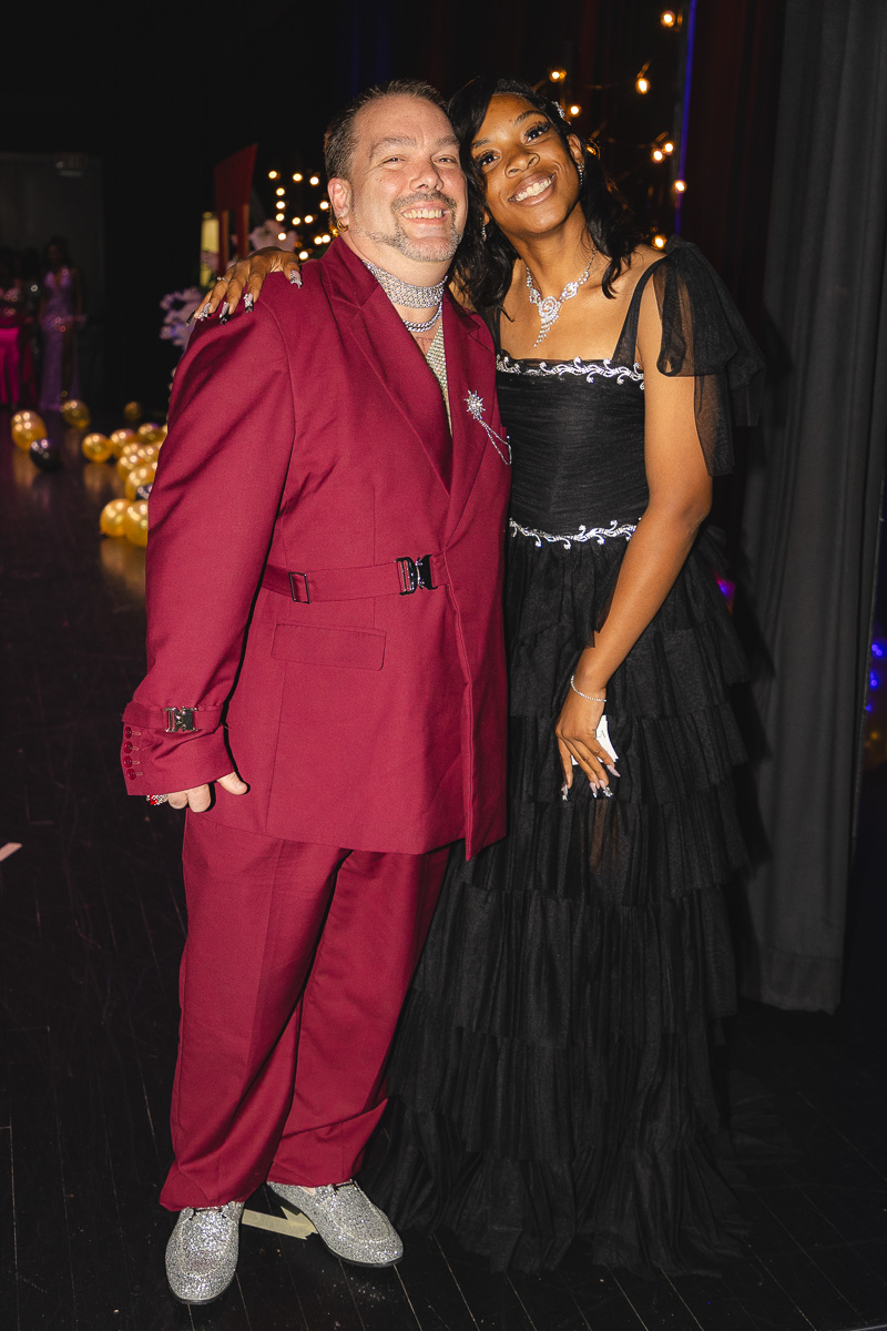 Smiling couple posing for a photo at a formal event: man in a burgundy suit with glittery shoes, woman in a black tiered gown with jewelry.