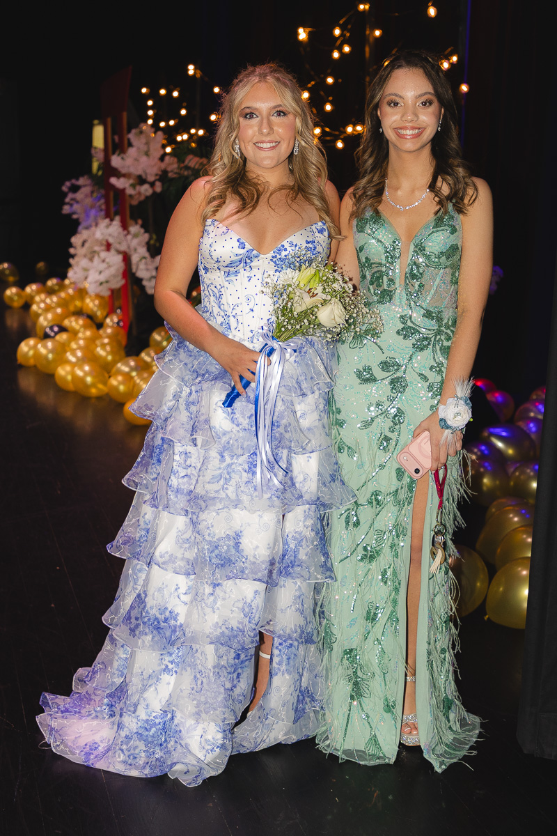 Two women in formal gowns posing backstage at a celebration; one in a blue-and-white tiered dress holding a bouquet, the other in a mint-green beaded gown.