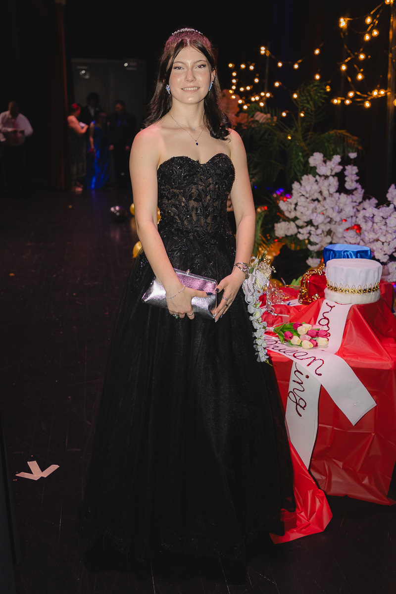 Young woman in a black strapless ball gown with a tiara, holding a pink clutch at a decorated formal event with string lights and gifts nearby.