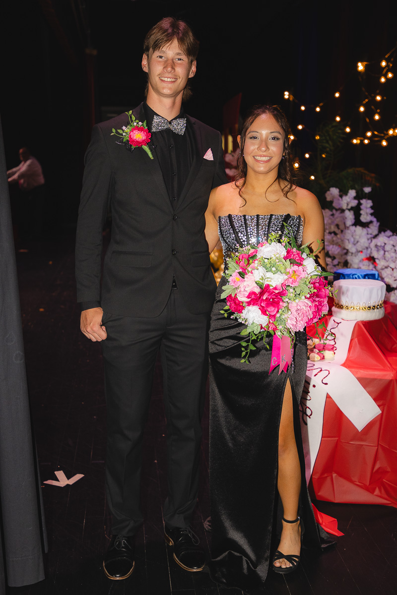 A young couple at a formal event posing together; man in a black suit with bow tie, woman in a strapless black gown holding a pink-and-white bouquet.