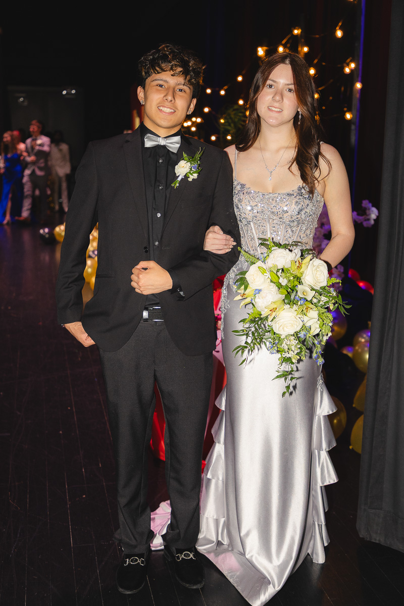 Couple in formal wear posing arm‑in‑arm on a stage; man in black suit with bow tie and boutonniere, woman in a silver beaded gown holding a white bouquet.