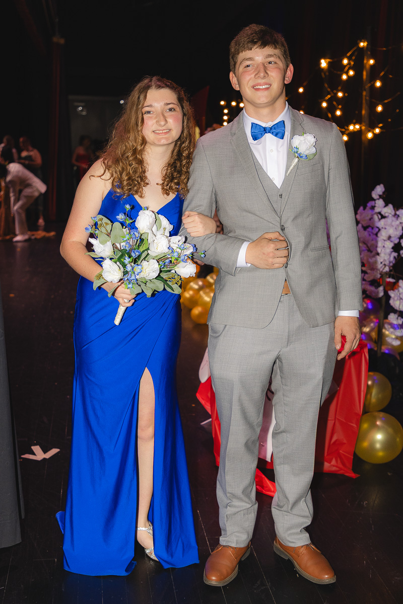 Two teens in formal wear pose arm-in-arm at a dance, the girl in a cobalt blue gown holding a white bouquet and the boy in a light gray suit with a blue bow tie.