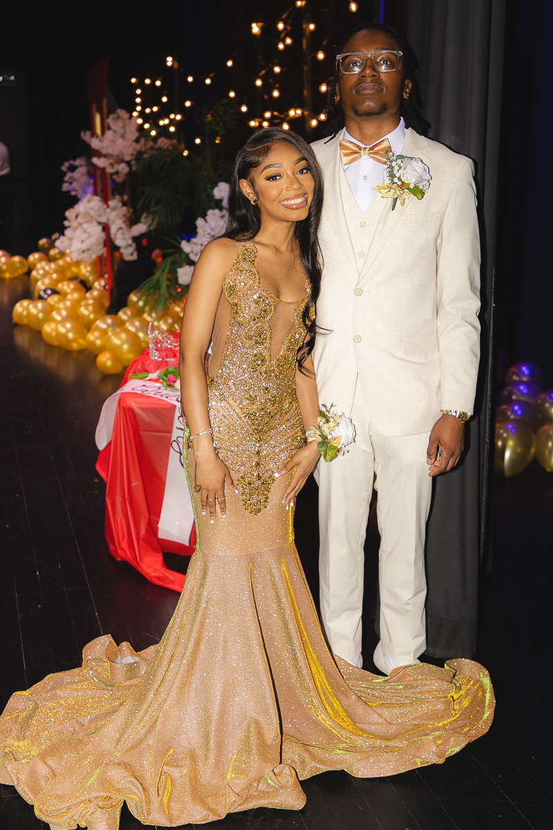 Couple posing at a formal event: woman in a gold sequin gown with a long train, man in a cream suit with bow tie.