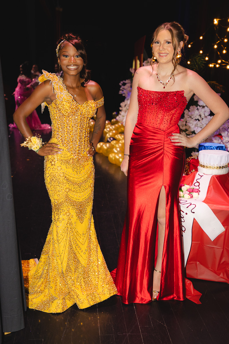 Two women in formal gowns pose on a stage; one in a yellow beaded dress, the other in a red satin gown, with balloons and a cake behind them.