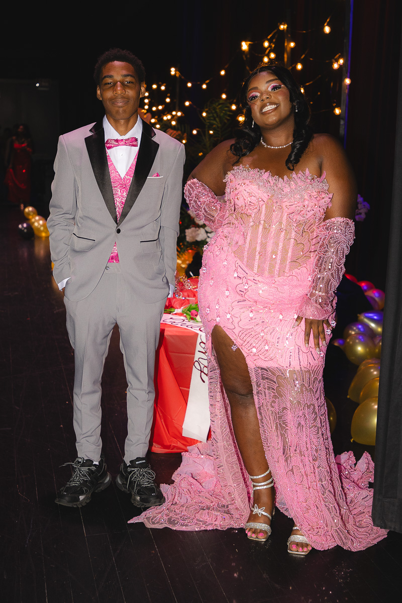 Two young people in formal attire posing at a party; woman in a pink lace gown and man in a gray suit with a pink vest smile at the camera.