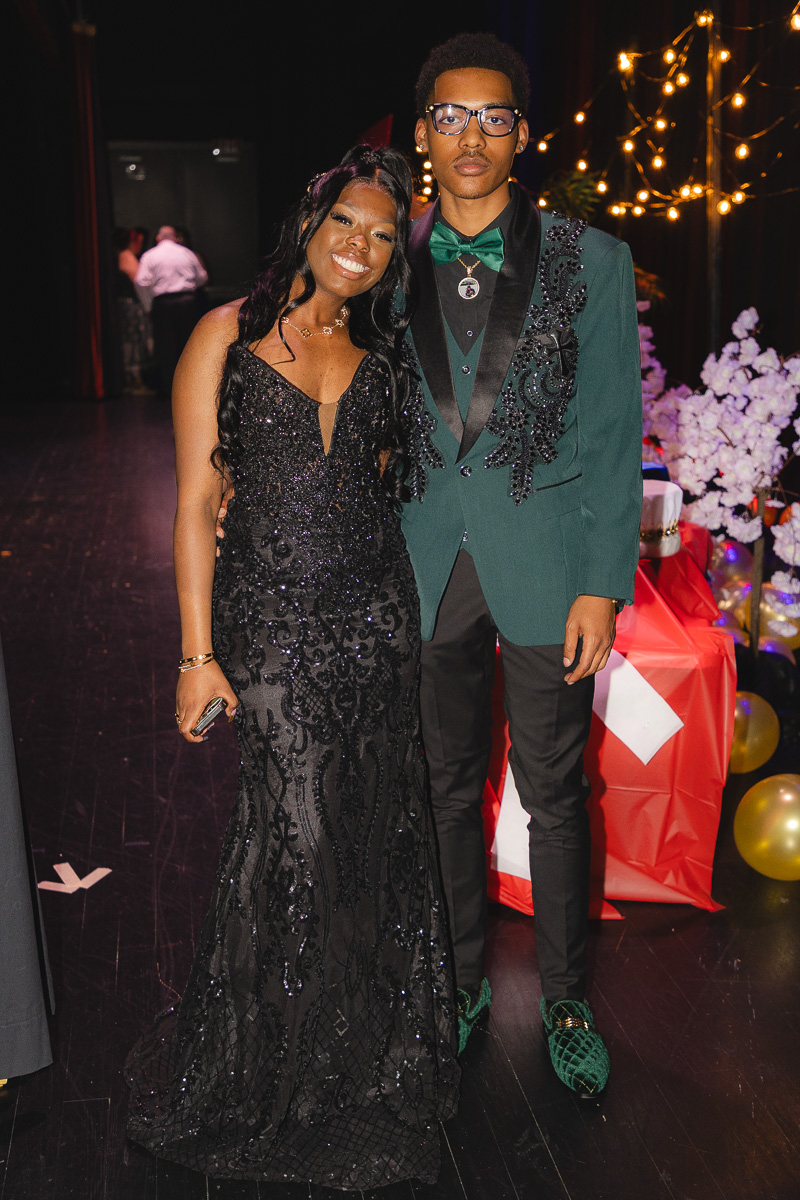 A smiling couple poses at a formal event: woman in a black sequin gown and man in a green tuxedo jacket with a bow tie and glasses.
