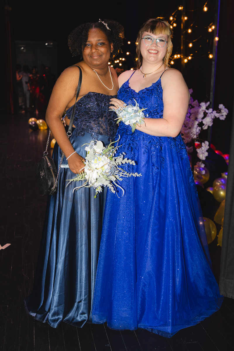 Two women in formal gowns pose together at a party, each holding a bouquet of white flowers and smiling under warm lights.",