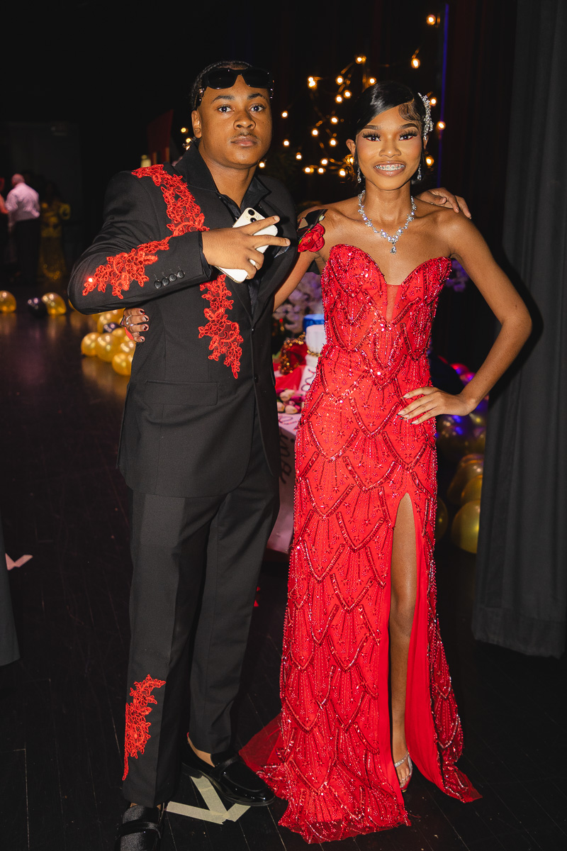 Couple posing for a photo at a formal event: woman in a red beaded gown with a high slit, man in a black suit with red embroidery.