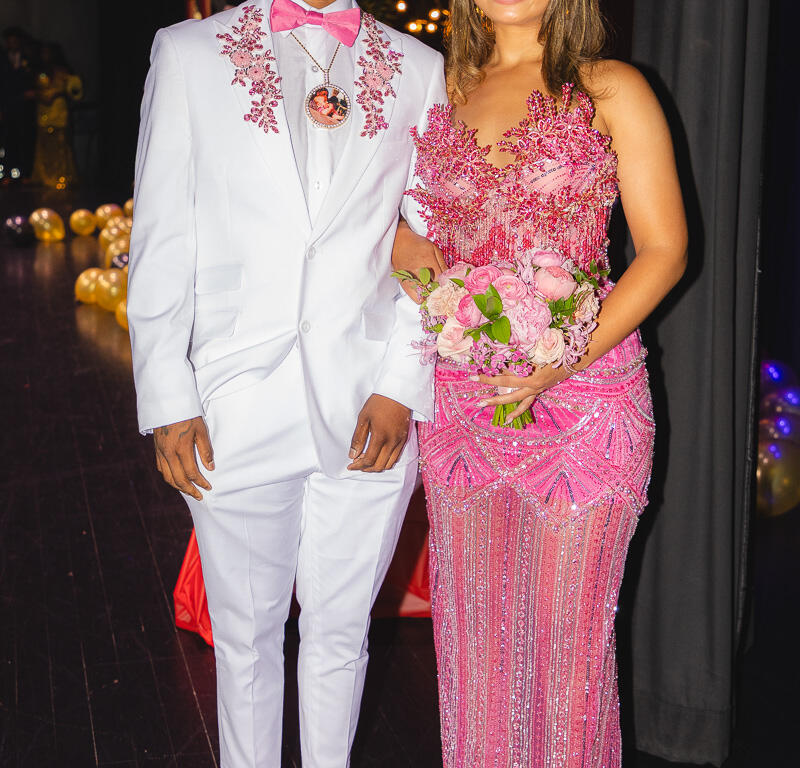 Couple dressed in formal attire posing together; man in a white suit with pink bow tie and pink loafers, woman in a pink embellished gown holding a bouquet.