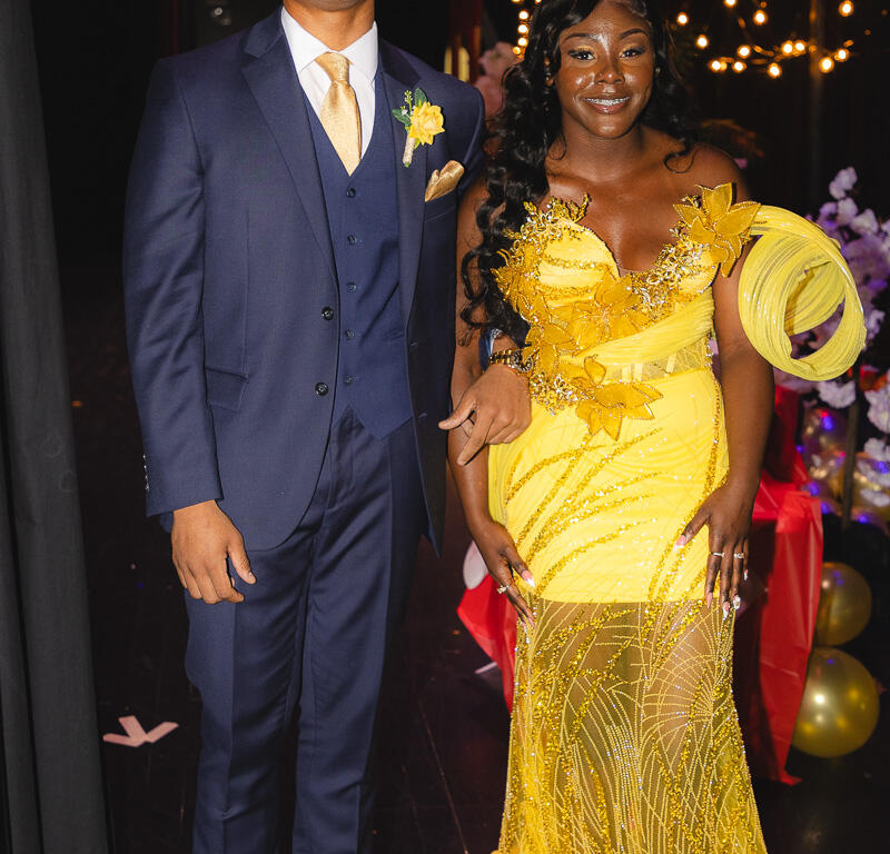 Young couple posing backstage in formal attire: man in navy suit with tan shoes, yellow tie, and boutonniere; woman in a yellow beaded evening gown with floral embellishments and a coordinated headpiece.